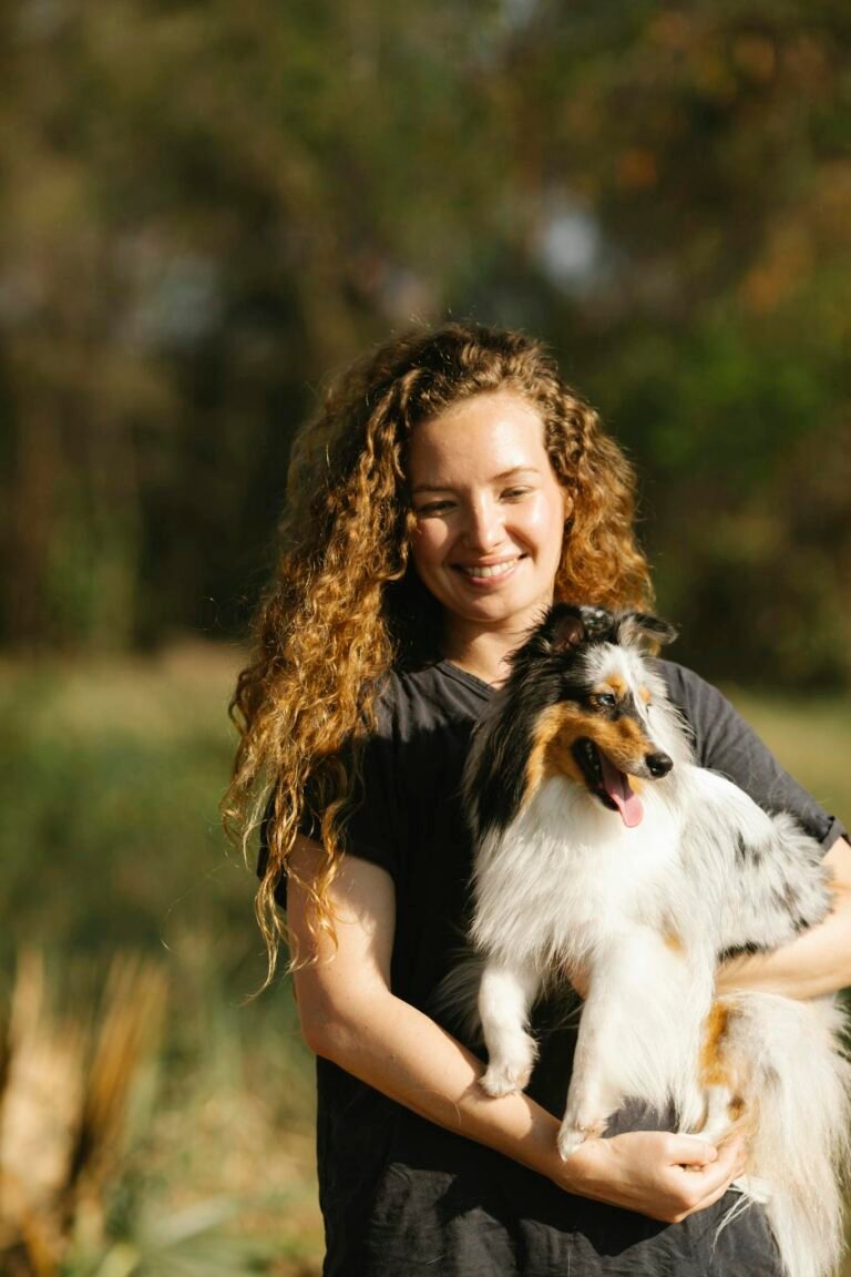 A cheerful woman outdoors holding a happy Shetland Sheepdog under the sunlight. Captures affection and companionship.