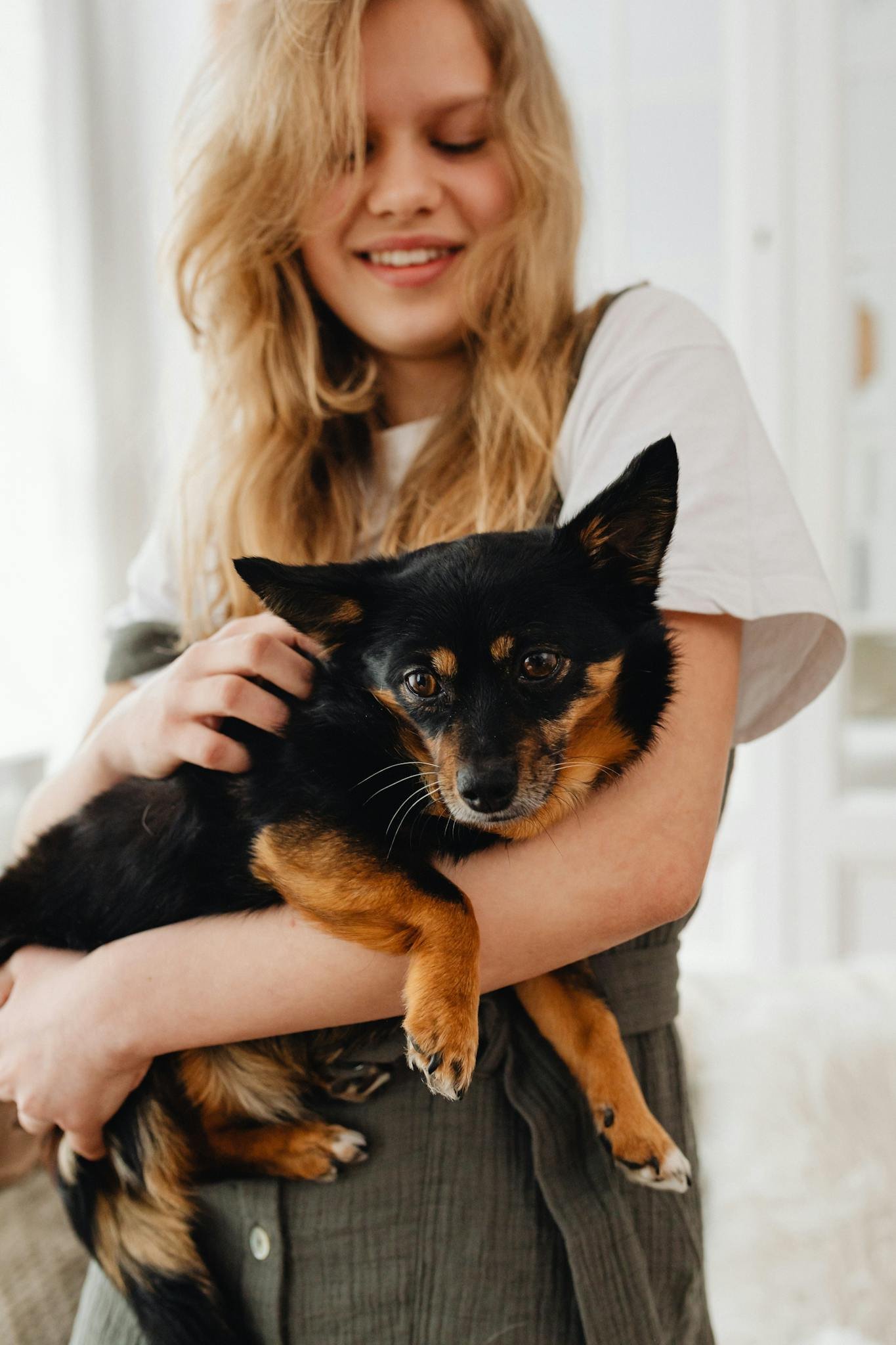 Smiling blonde girl holding a cute black dog indoors. Loving pet moment.